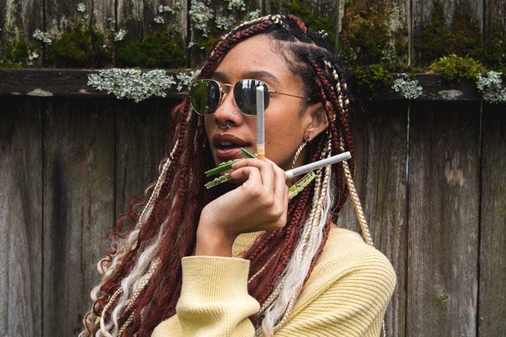 A woman with colorful braided dreadlocks, wearing sunglasses and holding multiple infused pre-roll joints, showcasing her love for smoking herb.