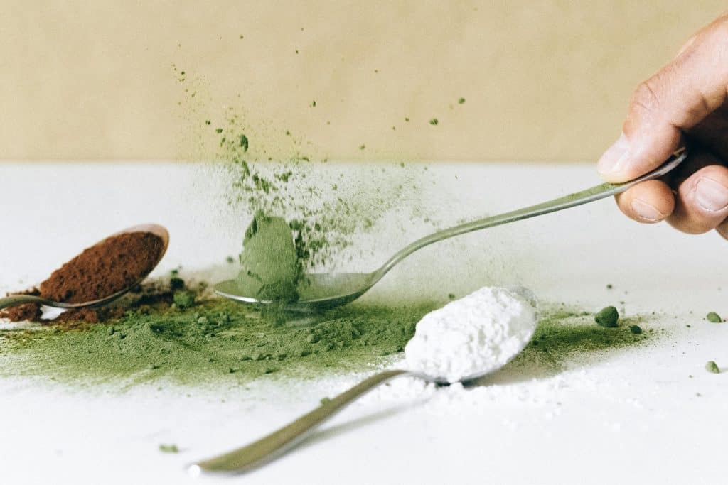 Close-up of green and white vape flavor powders being scooped with metal spoons, highlighting ingredients used in e-liquid flavoring.