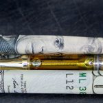 $100 bill, vape pen, and rolled-up dollar bills on dark surface - vaping, vape deals, and financial savings.