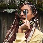 A woman with colorful braided dreadlocks, wearing sunglasses and holding multiple infused pre-roll joints, showcasing her love for smoking herb.